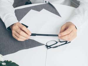 Close-up of hands holding a pen with papers and eyeglasses on a white table.